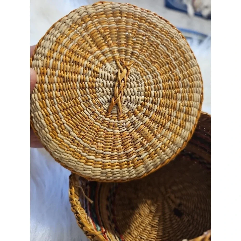 The image displays a‎ small, round, hand-woven basket with a lid, typically made